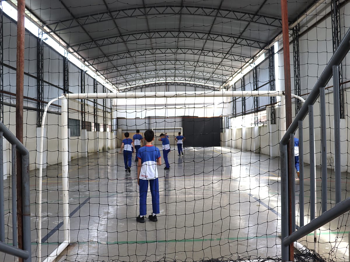 Alunos jogando bola na quadra coberta, vistos através da rede do gol